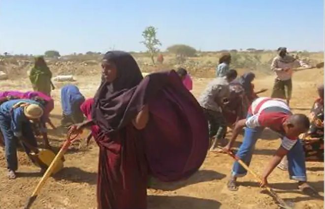 Woman in a dark shawl standing among workers hoeing the catchment floor