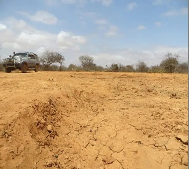 Shallow eroded pan of dry red earth with a vehicle parked at the far rim